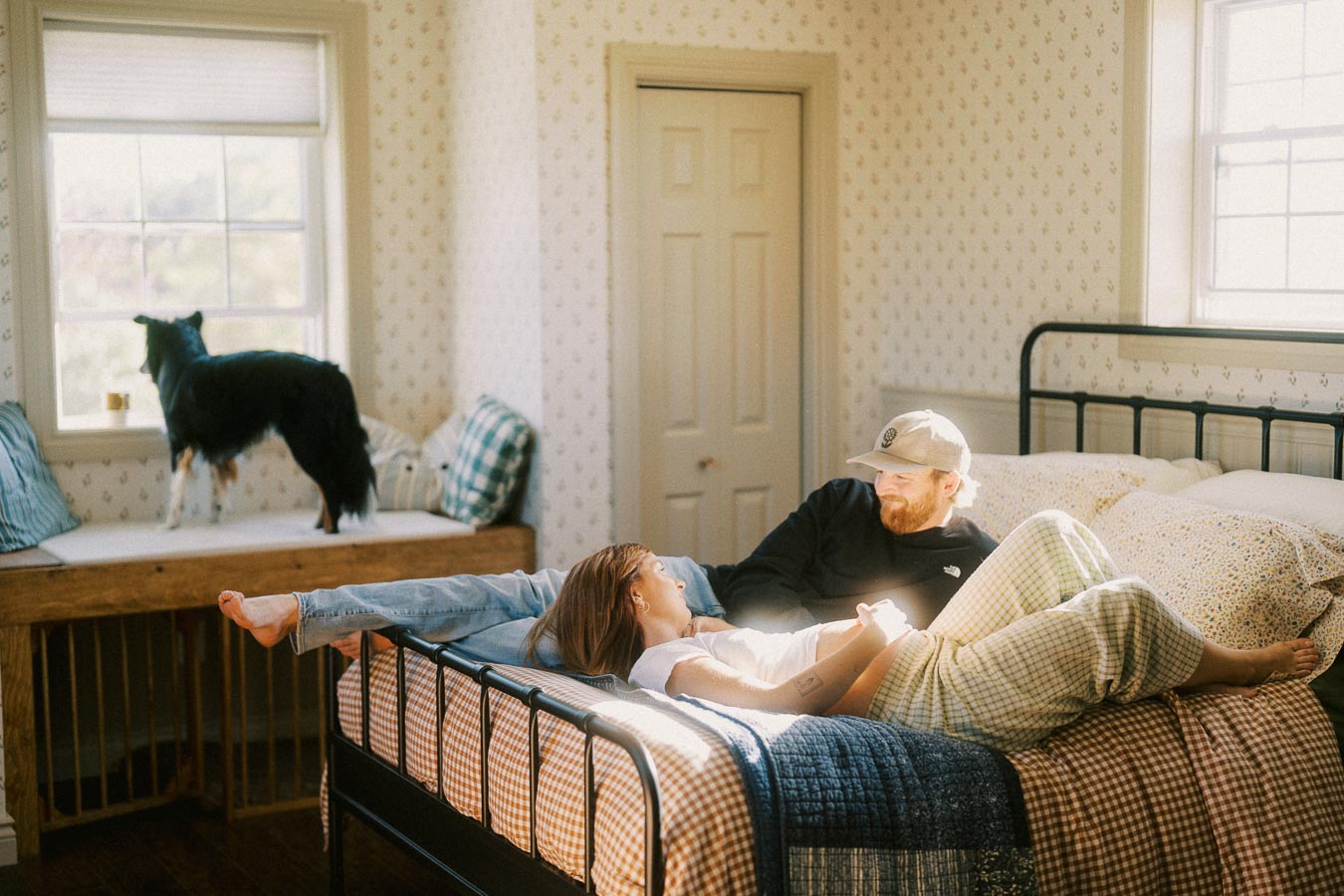 A couple relaxing on a sunlit bed in a cozy bedroom with a patterned wallpaper, while a dog stands by the window seat looking outside.