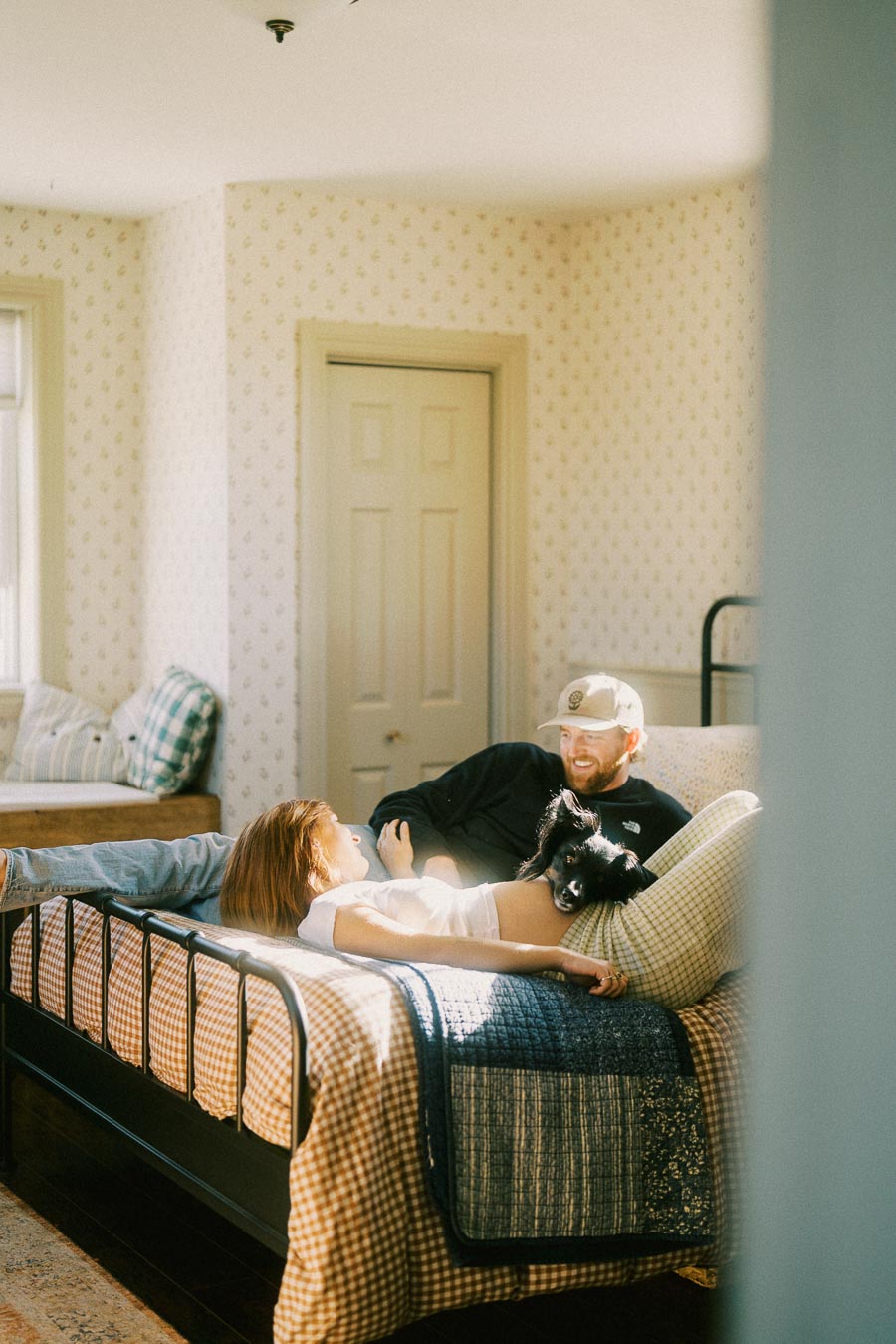 A couple relaxing on a bed in a sunlit bedroom, with a small black dog resting on the woman's stomach, creating a cozy and intimate atmosphere.