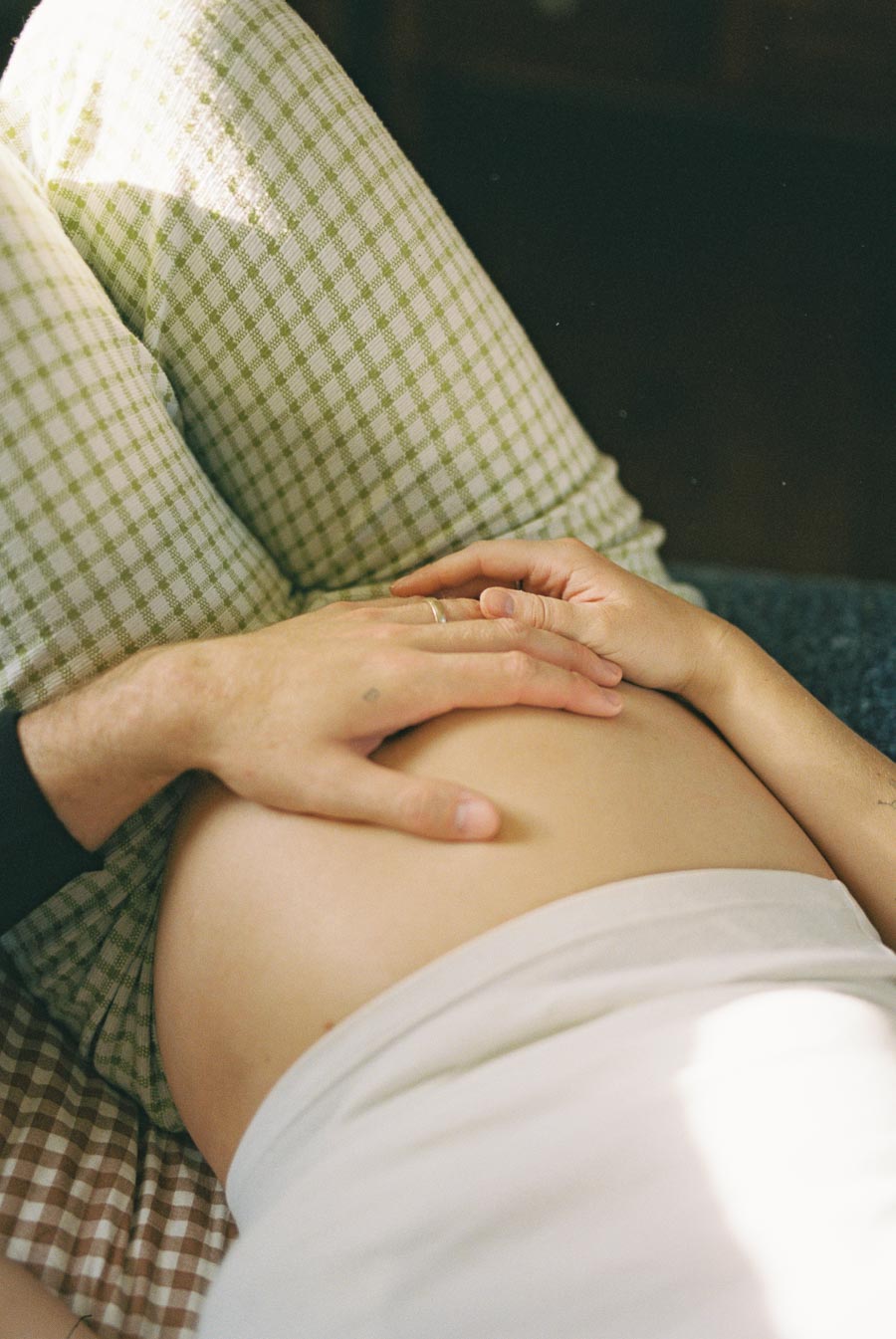 A close-up of a pregnant belly with two hands gently resting on it, conveying love and anticipation. The person is wearing patterned pants, suggesting a relaxed, intimate setting.