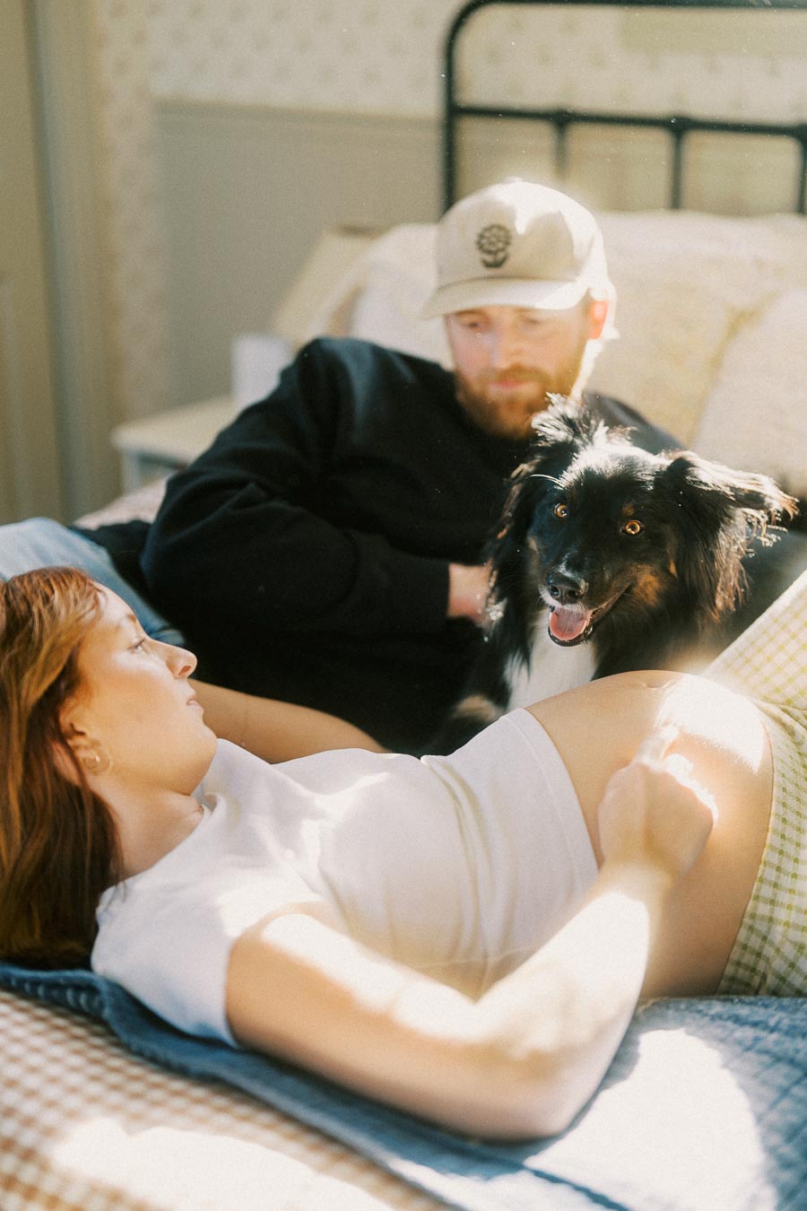 A couple relaxing on a bed with their happy black and white dog in between, sunlight streaming through a window.