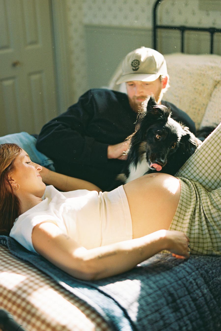 A pregnant woman lying on a bed with a dog and a man, enjoying a relaxed moment in a sunlit bedroom.