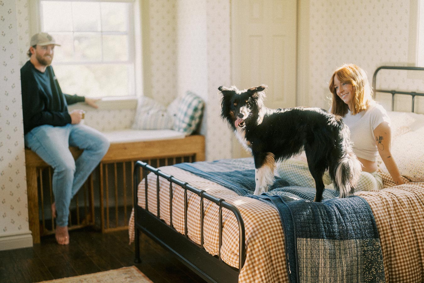 Cozy bedroom scene featuring a black and white dog standing on a bed with a blue quilt, while a smiling woman sits nearby. A man in casual attire sits at a window seat with natural light streaming in, creating a warm, inviting atmosphere.