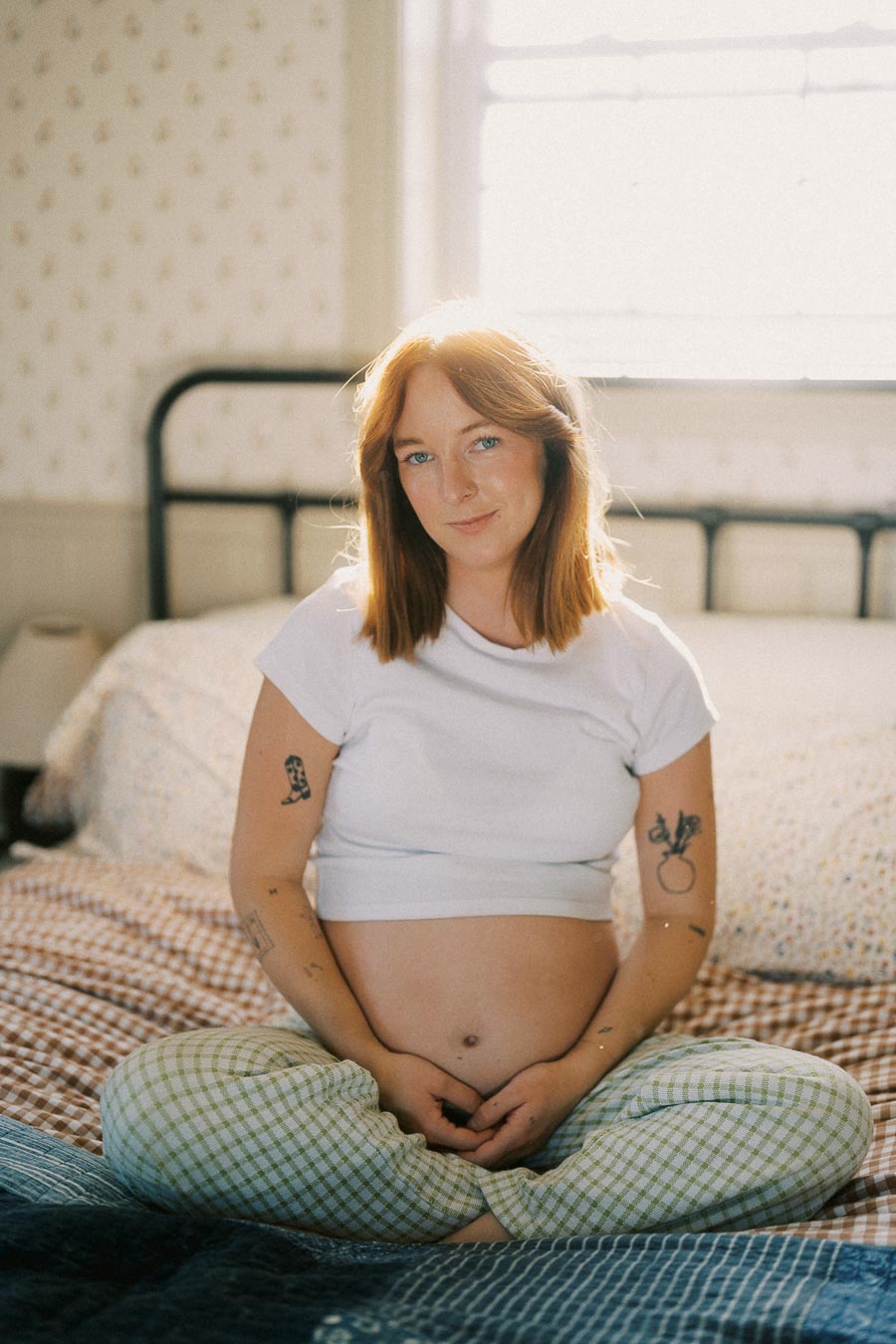 Pregnant woman sitting cross-legged on a bed, wearing a white shirt and green plaid pants, with tattoos visible on her arms, in a softly lit bedroom.