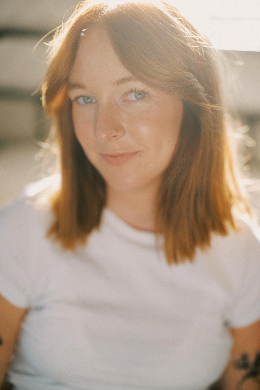 A woman with shoulder-length hair smiling softly in a bright, naturally lit setting, wearing a white t-shirt and a nose ring.