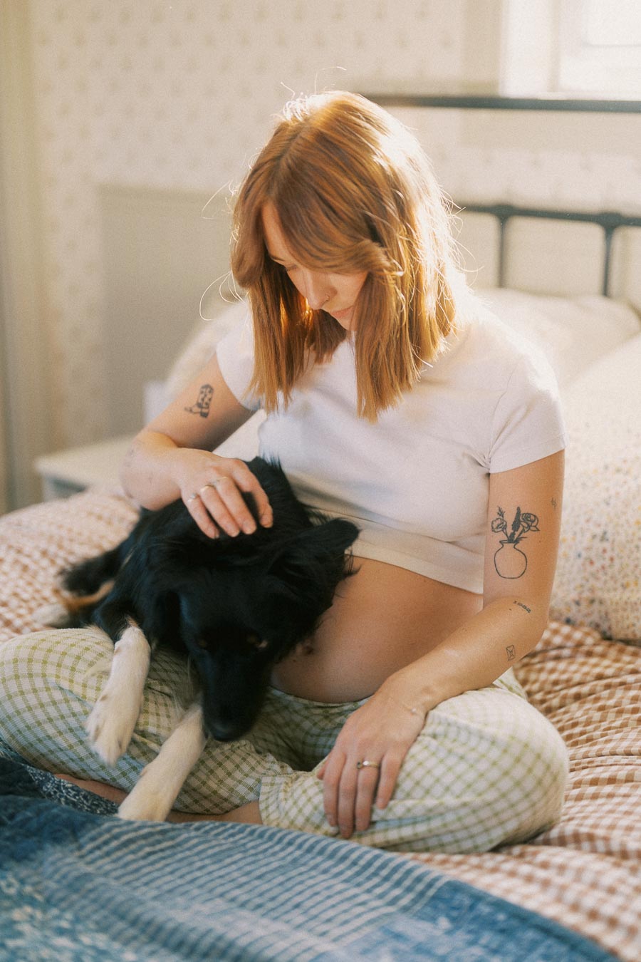 Pregnant woman sitting on bed with black dog, gently caressing its head. She is wearing a white t-shirt and plaid pajama pants, showcasing tattoos on her arms in a cozy bedroom setting.