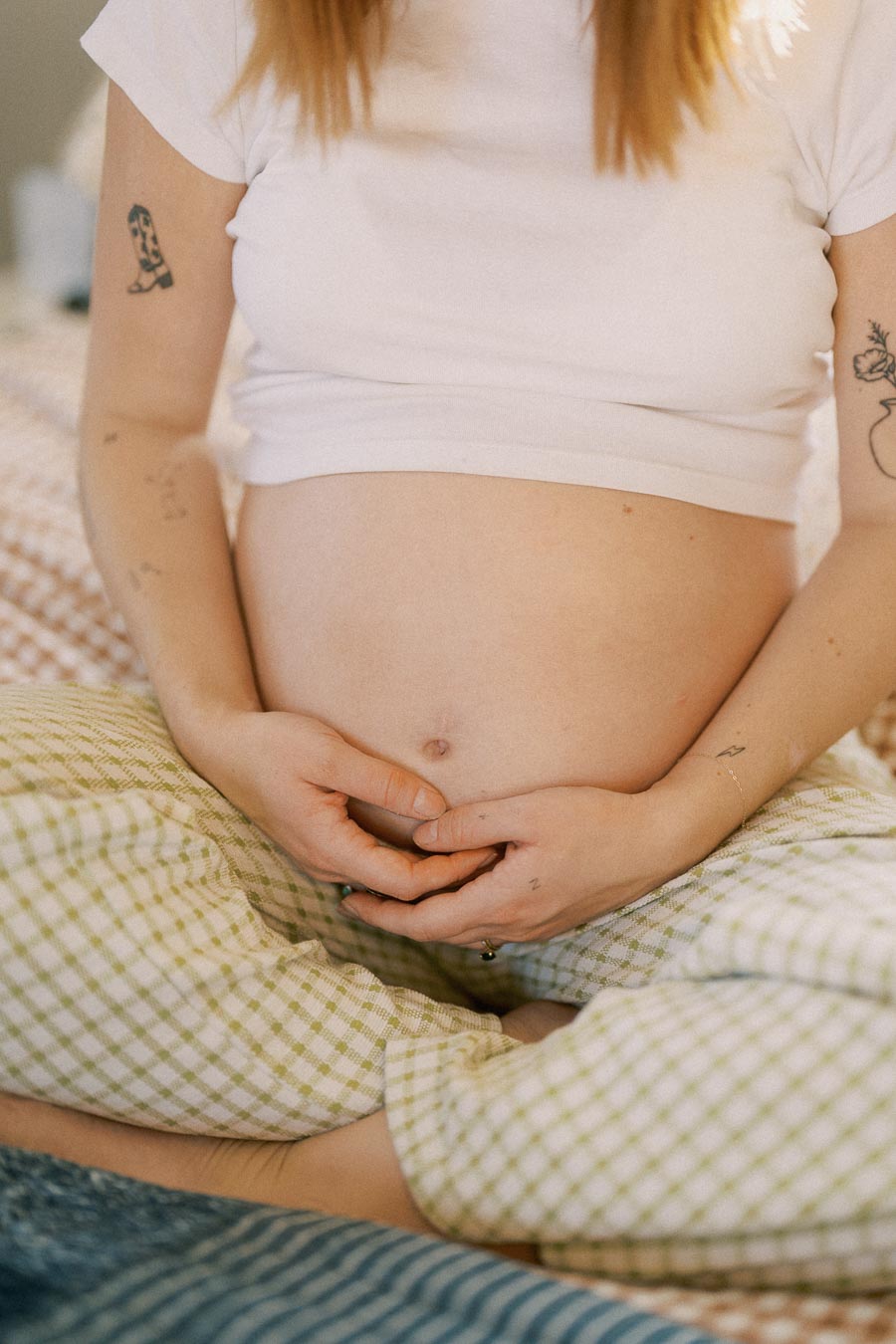 A pregnant woman sitting cross-legged on a bed, gently cradling her belly with her hands, wearing a white cropped shirt and checkered pajama pants.