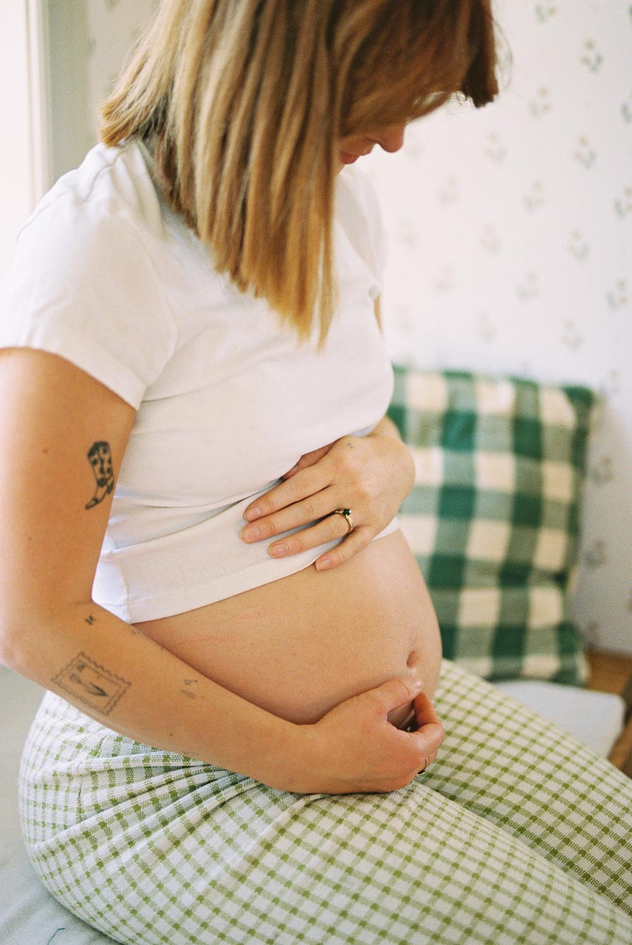 Pregnant woman gently holding her belly while sitting on a bed, wearing a white t-shirt and checkered pants.