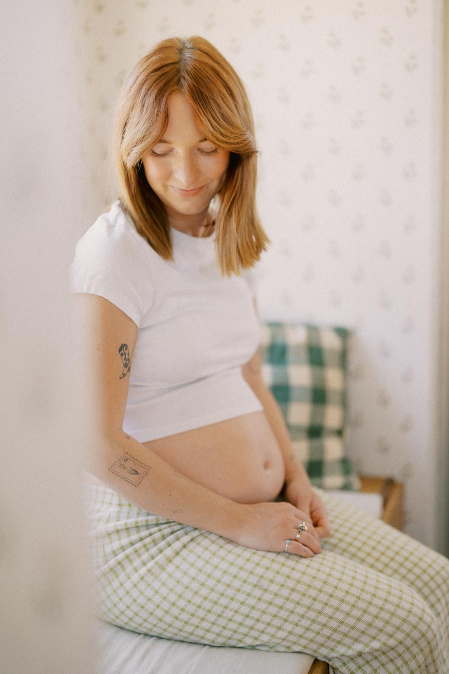 Smiling pregnant woman sitting on a bed, wearing a white shirt and checkered pants, looking down with a peaceful expression.