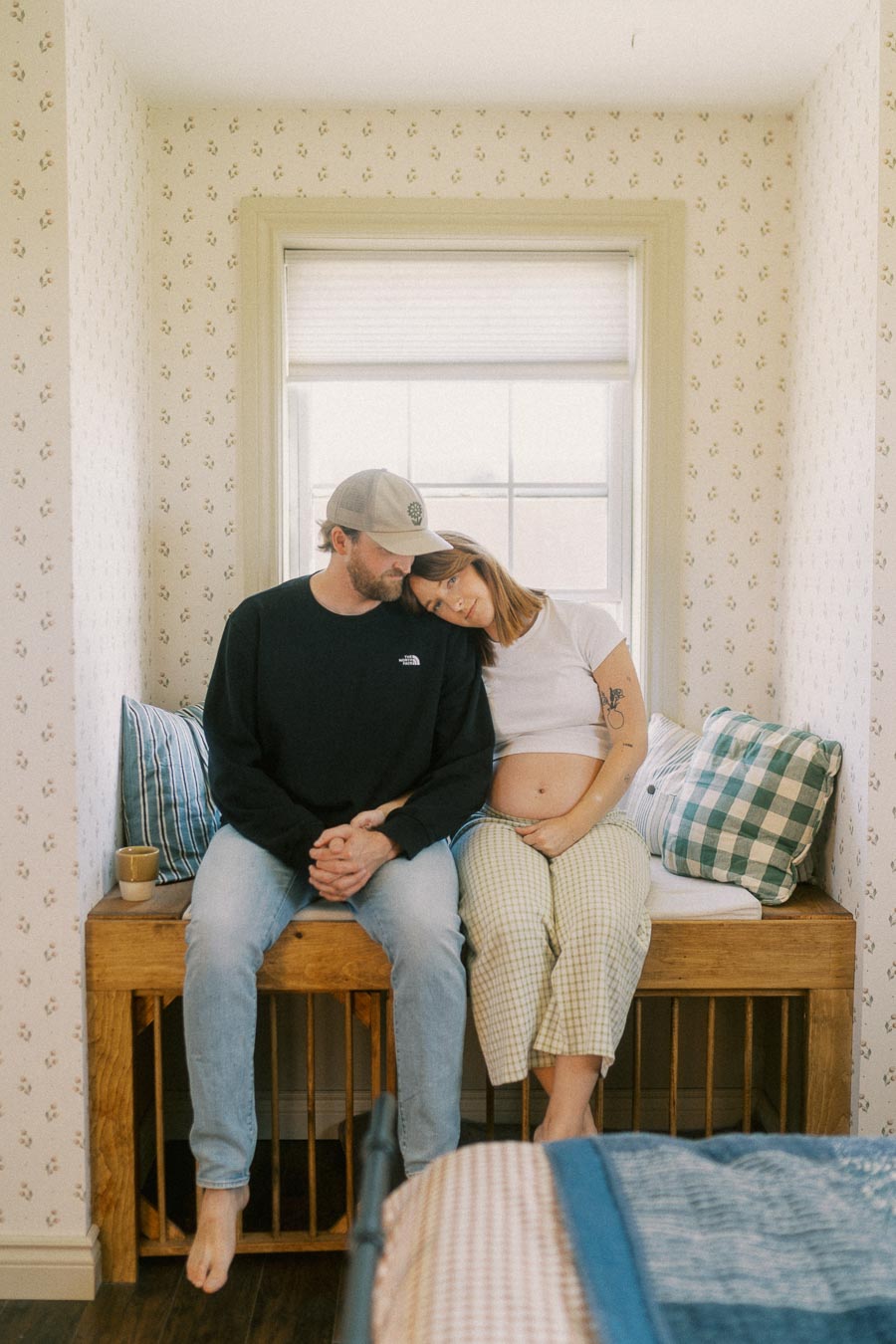 Couple sitting together in a cozy room, expecting a baby. Woman with a visible baby bump leaning on the man. Bright window, patterned wallpaper, and striped cushions enhance the warm setting.