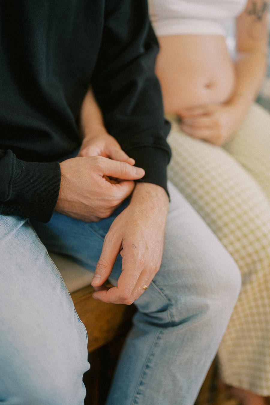 A couple sitting together, with the focus on their intertwined hands, conveying warmth and connection. One person is wearing a dark sweatshirt and jeans, while the other is in a checkered outfit, possibly indicating a casual or intimate setting.