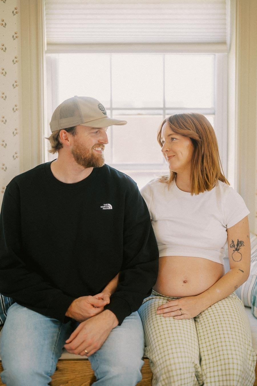 A couple sitting together on a bench, looking at each other with smiles. The woman is pregnant, wearing a white crop top and checkered pants, while the man is wearing a cap, a black sweatshirt, and jeans. They're indoors with a window in the background.