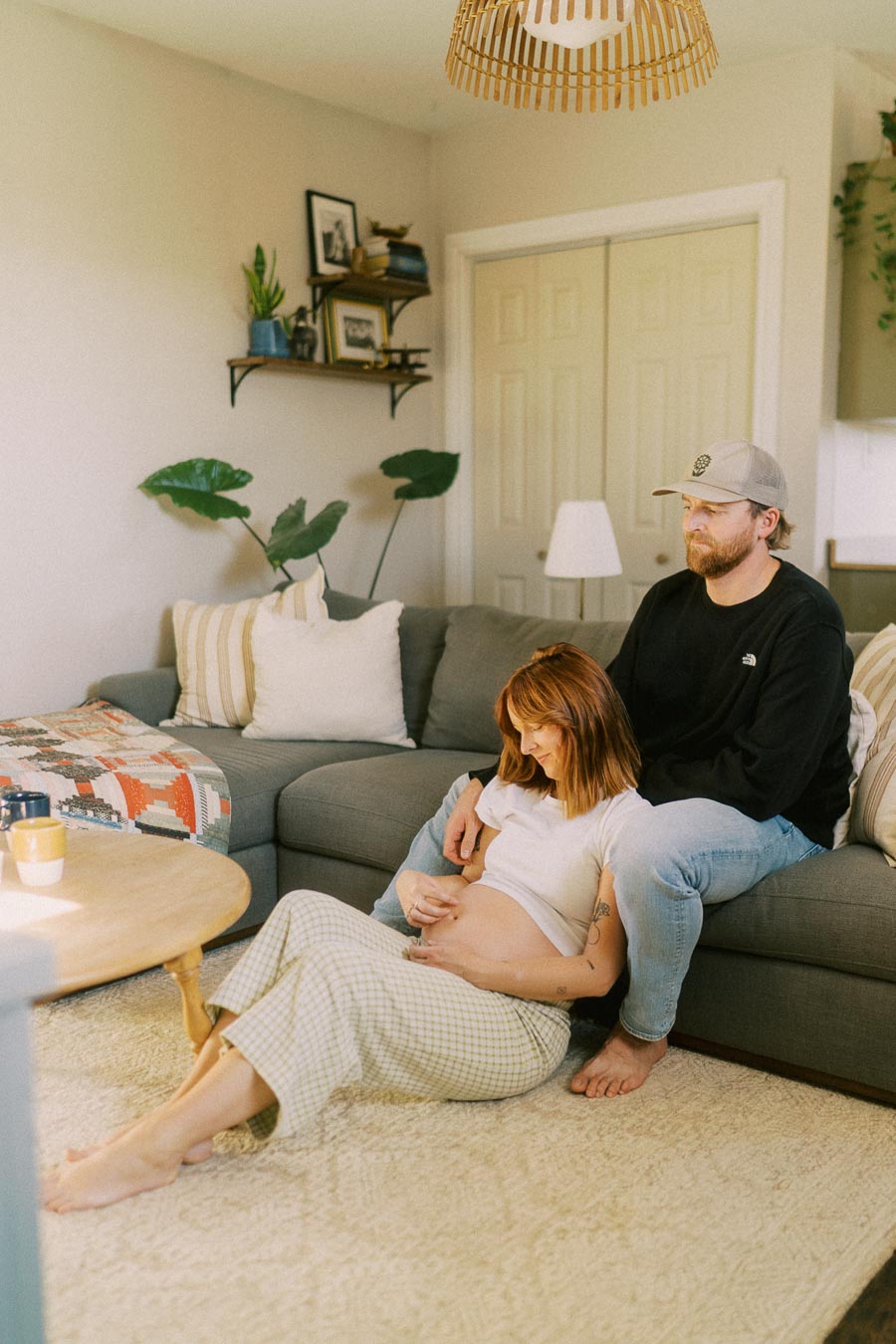 A couple sitting in a cozy living room, with the woman resting against the man on a gray sofa. She is gently cradling her pregnant belly, while the man lovingly supports her from behind. The room features modern decor, including a coffee table, indoor plants, and a patterned throw blanket.