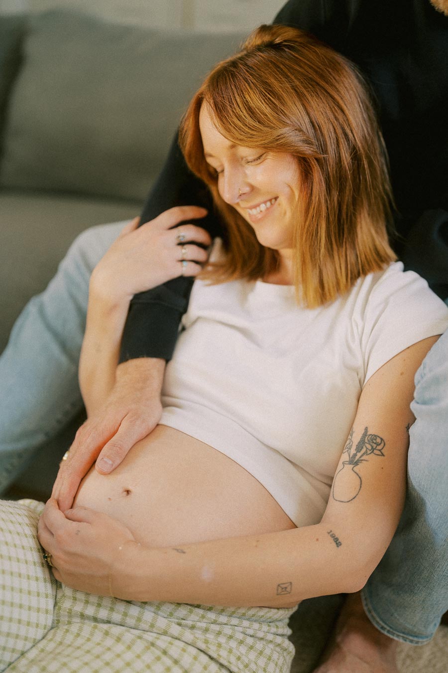 A smiling pregnant woman sitting on a couch, gently holding her belly with both hands, accompanied by a partner's supportive embrace. The scene conveys warmth, affection, and the joy of expecting a baby.