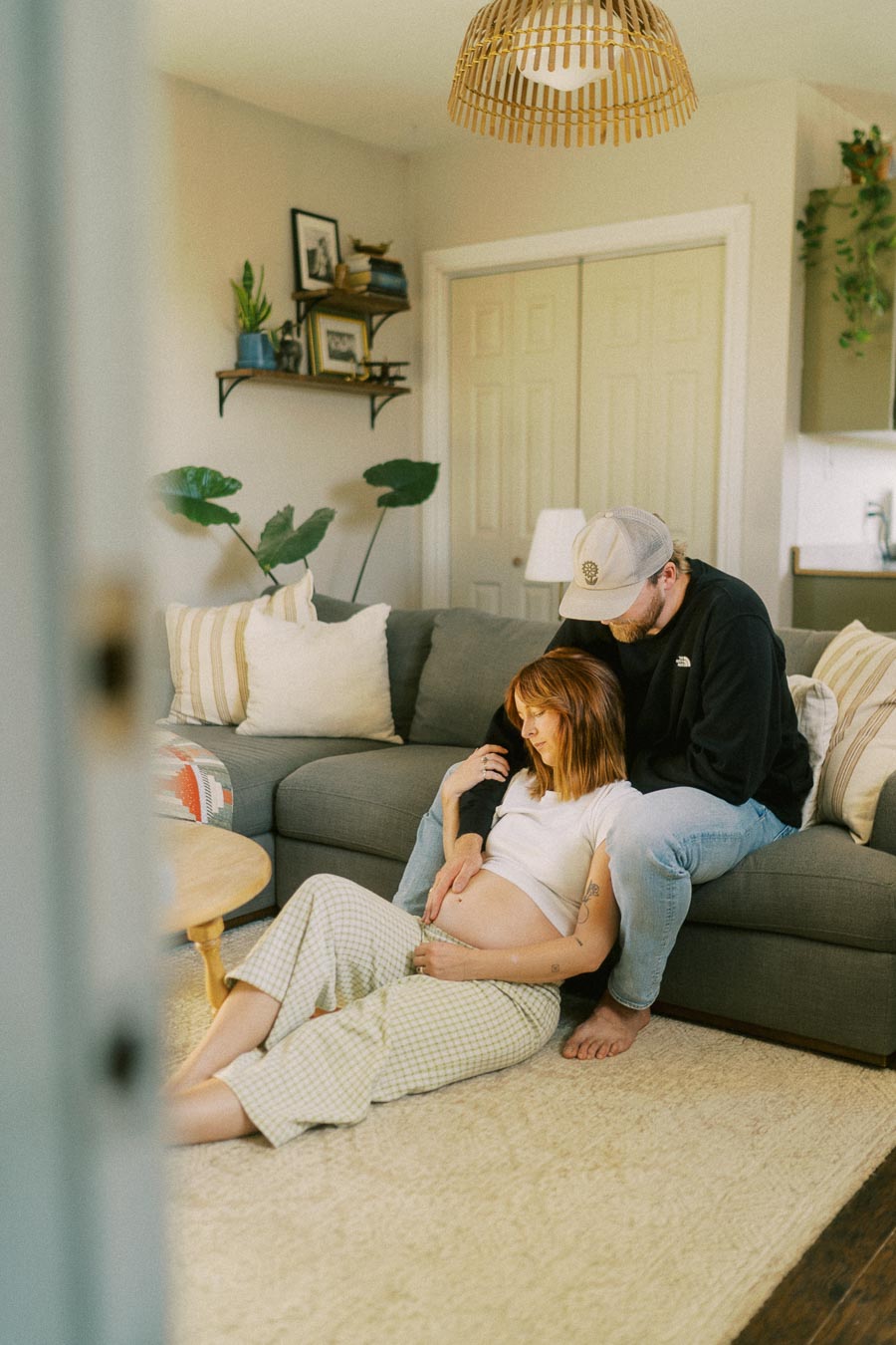 A couple sitting on the floor of a cozy living room, with a woman showcasing her pregnant belly and a man gently embracing her from behind, surrounded by soft furnishings and indoor plants.