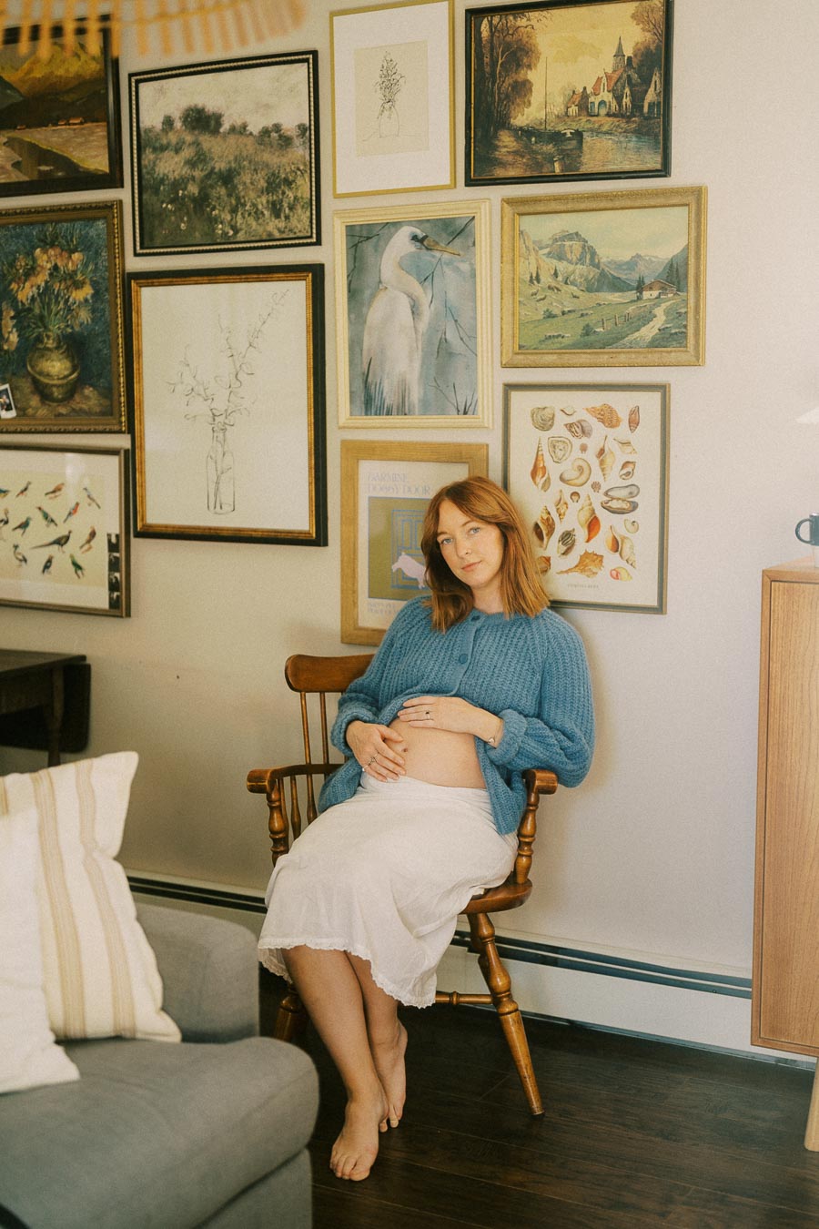 Pregnant woman in a cozy room with a gallery wall, sitting on a wooden chair, wearing a blue sweater and white skirt, holding her belly and surrounded by framed art.