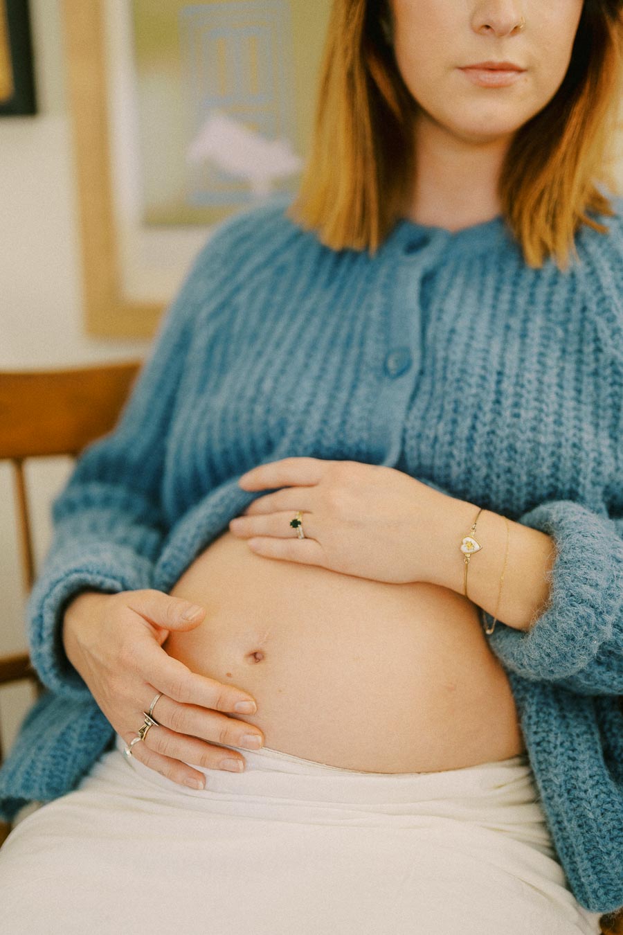 Pregnant woman gently resting hands on her belly while wearing a cozy blue sweater, sitting on a wooden chair in a warmly lit room.
