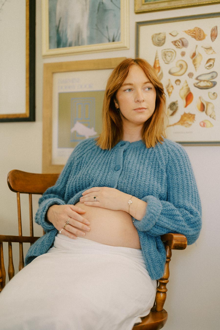 Pregnant woman in a blue sweater sitting in a wooden chair, holding her belly, surrounded by framed artwork on the wall.