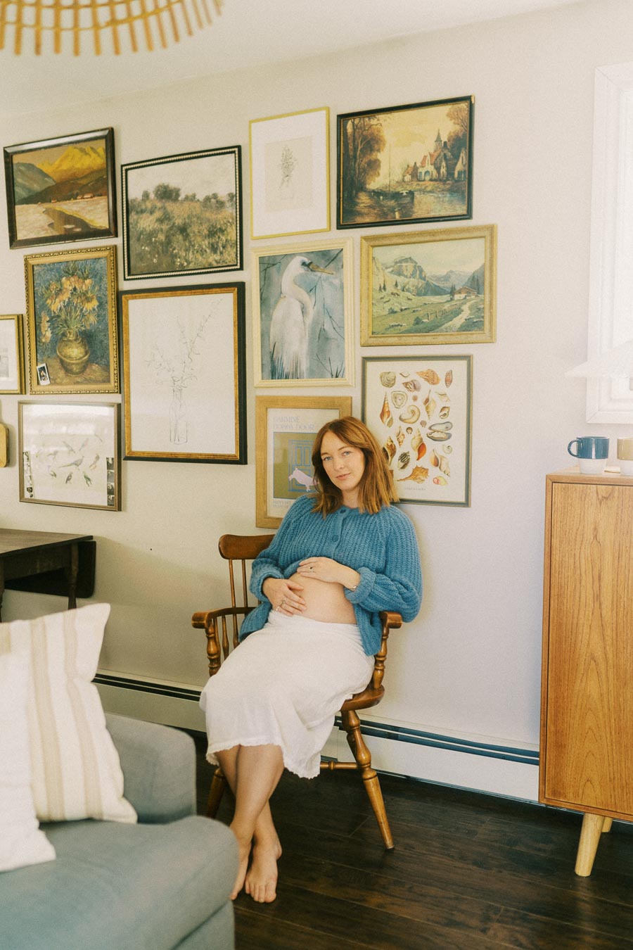 Pregnant woman in cozy living room with gallery wall, wearing a blue cardigan, sitting on a wooden chair, and holding her belly.