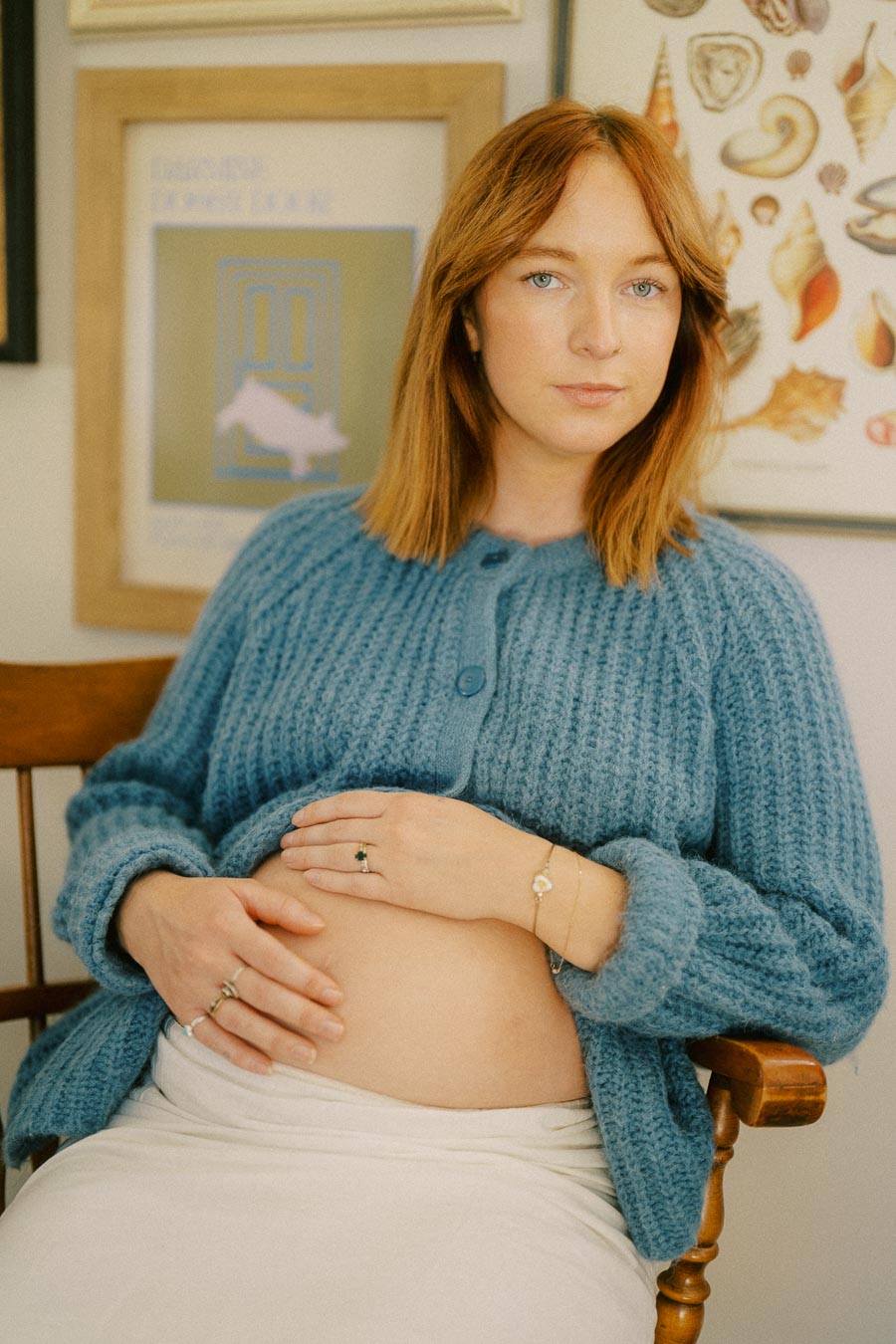 Pregnant woman sitting in a chair, wearing a blue knitted sweater, gently cradling her belly against a background of framed art.