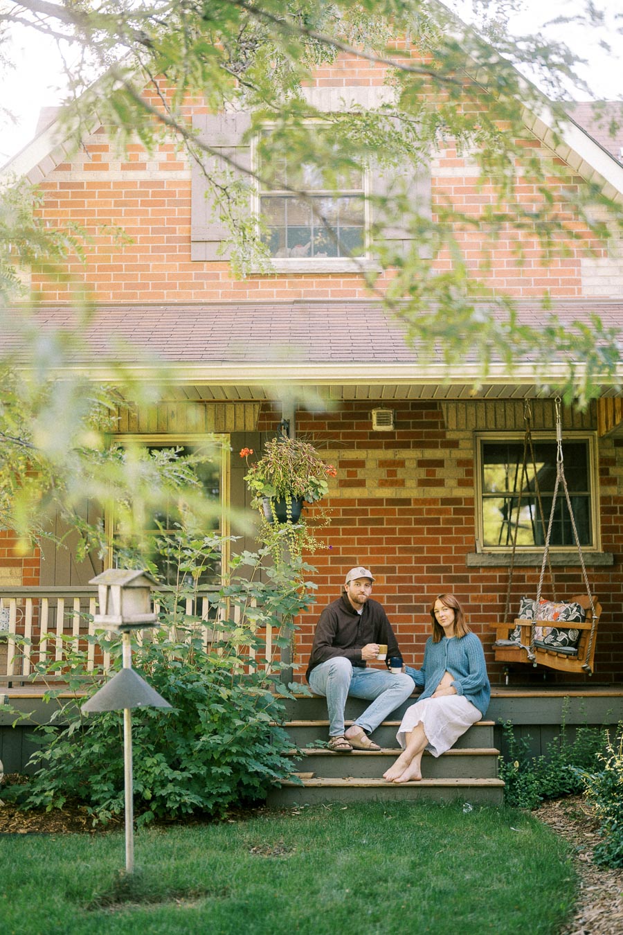 Couple sitting on porch steps of a brick house, surrounded by greenery, enjoying a peaceful morning with coffee mugs.
