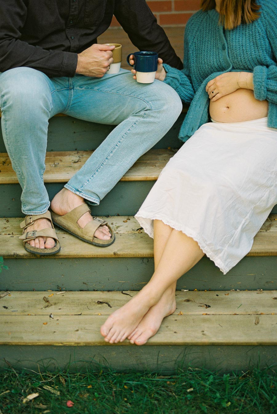 Couple sitting on wooden steps, holding mugs, with a focus on the woman's pregnant belly and relaxed barefoot pose, wearing casual attire.