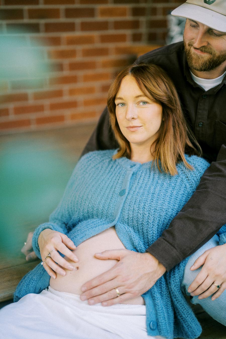 A pregnant woman with a blue sweater is sitting outdoors, being lovingly embraced by a man wearing a cap and dark shirt. Her hands and his are gently placed on her belly. Warm, intimate, and expectant moment against a brick wall background.