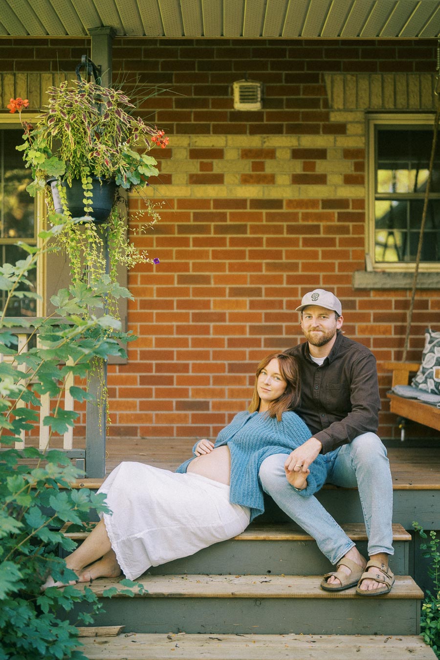 A pregnant woman in a blue sweater and white skirt sits on porch steps, leaning on a man in a cap and brown shirt, with a brick wall and hanging plants in the background.