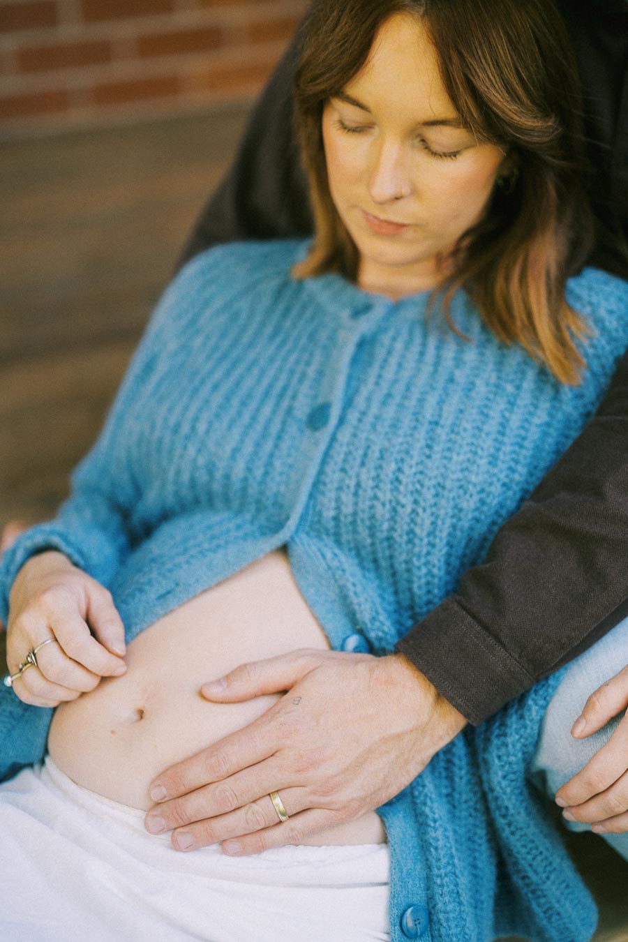 A pregnant woman wearing a blue sweater sits with her partner's hands gently resting on her bare belly, symbolizing warmth and connection during pregnancy.