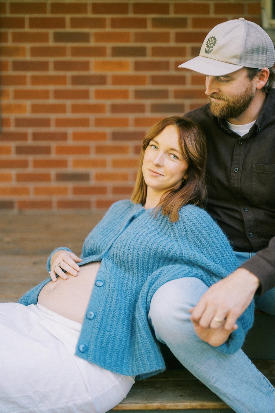 A pregnant woman in a blue cardigan and white pants lies against a man's legs on wooden steps in front of a brick wall, while they hold hands, conveying a sense of warmth and anticipation.