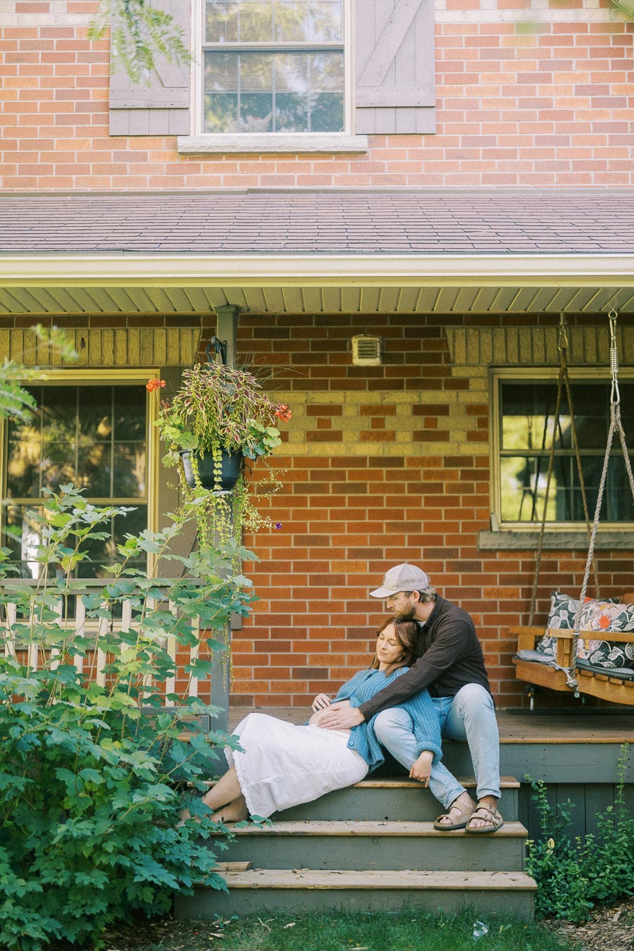 A couple sitting on the porch steps of a brick house, surrounded by greenery, with a hanging flower pot nearby. The man gently embraces the woman, creating a peaceful and intimate scene. Perfect for themes of home, love, and relaxation.