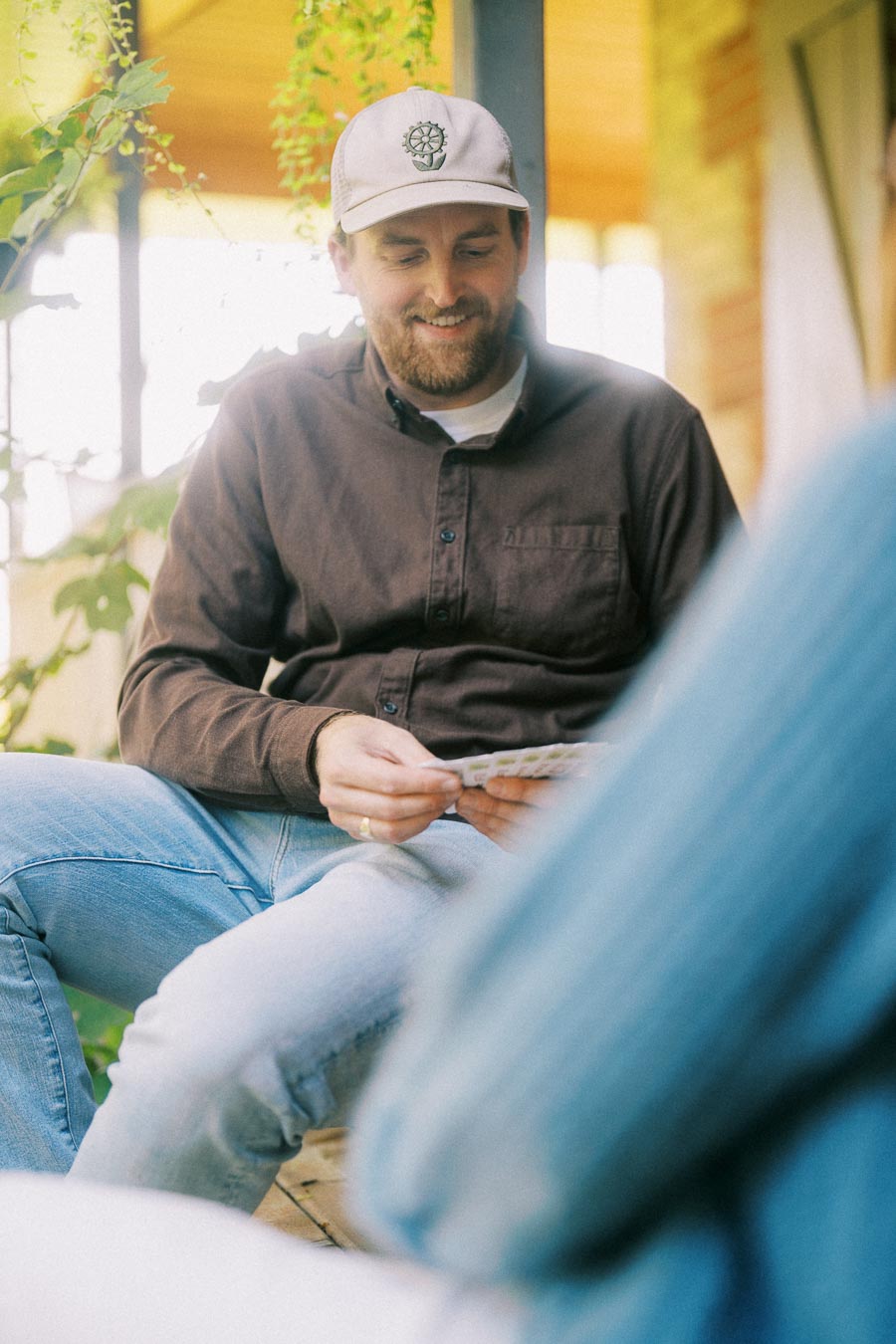 A man wearing a cap and a brown shirt smiles while sitting on a porch, holding a deck of cards. Background features greenery and soft natural light, creating a relaxed atmosphere.
