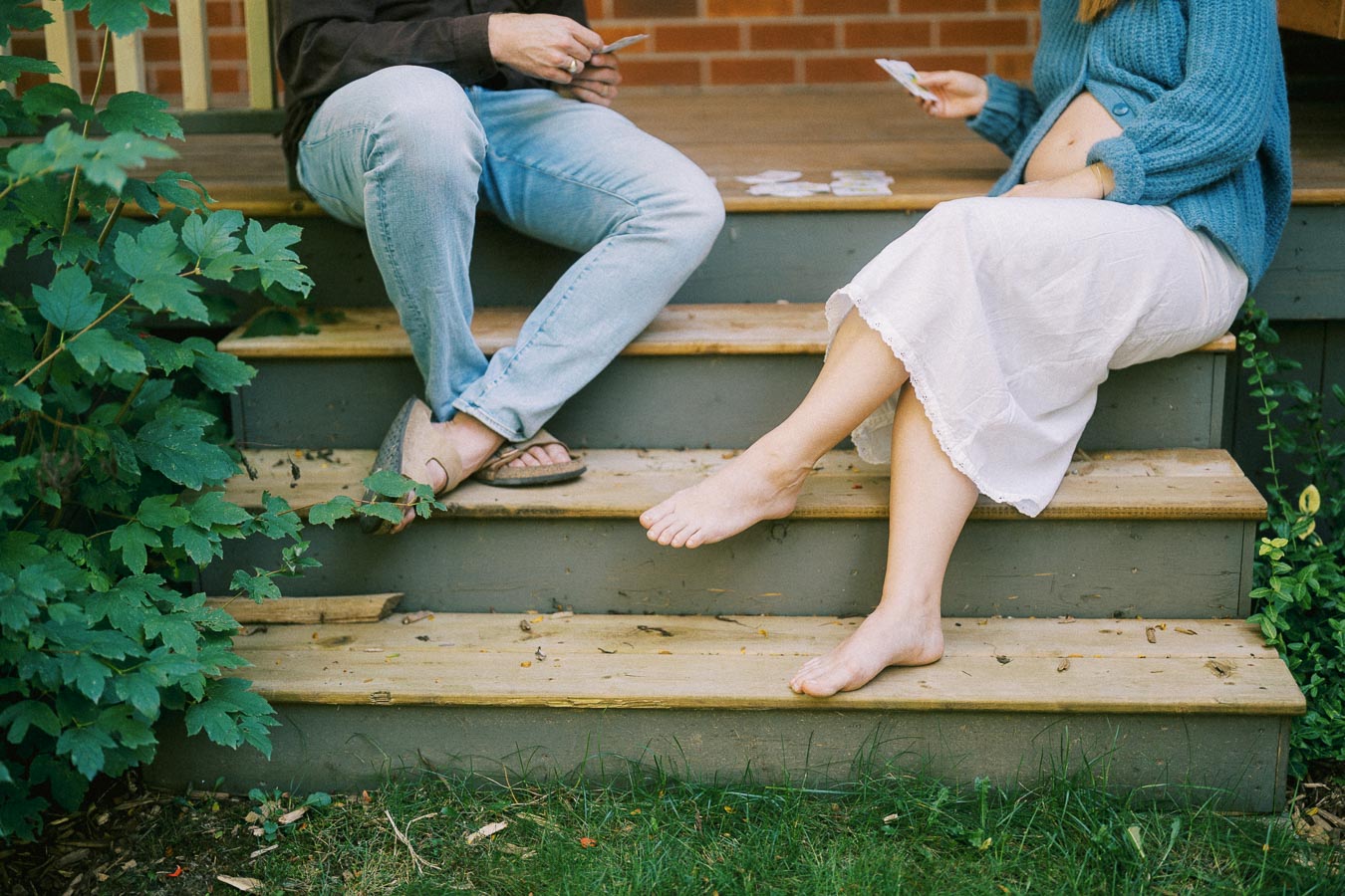 Couple sitting on wooden porch steps, playing cards; woman in blue sweater and white skirt, man in jeans and casual sandals, surrounded by greenery.