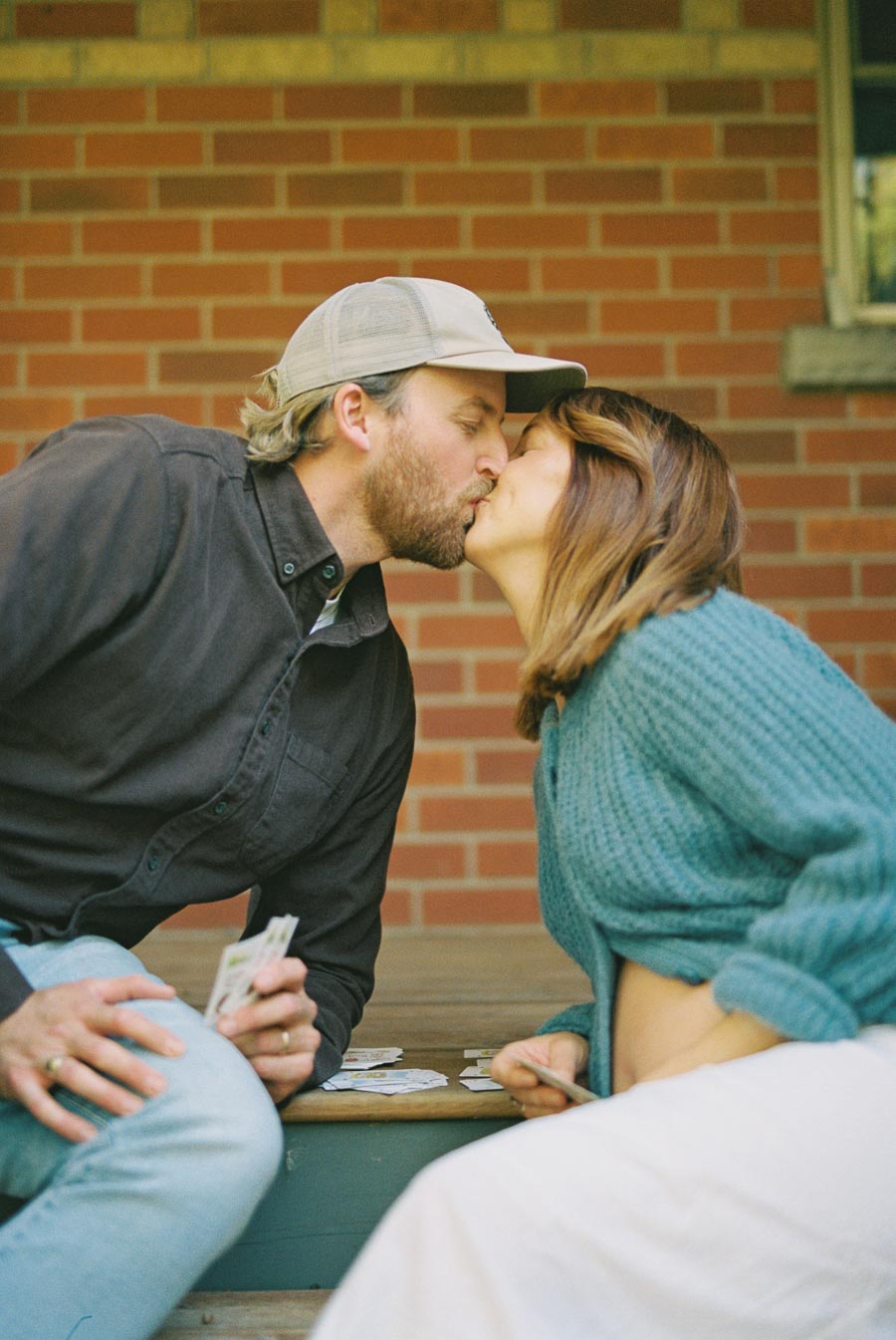 A couple shares a kiss while sitting on a wooden bench in front of a brick wall, each holding playing cards. They appear to be enjoying a relaxed, intimate moment.
