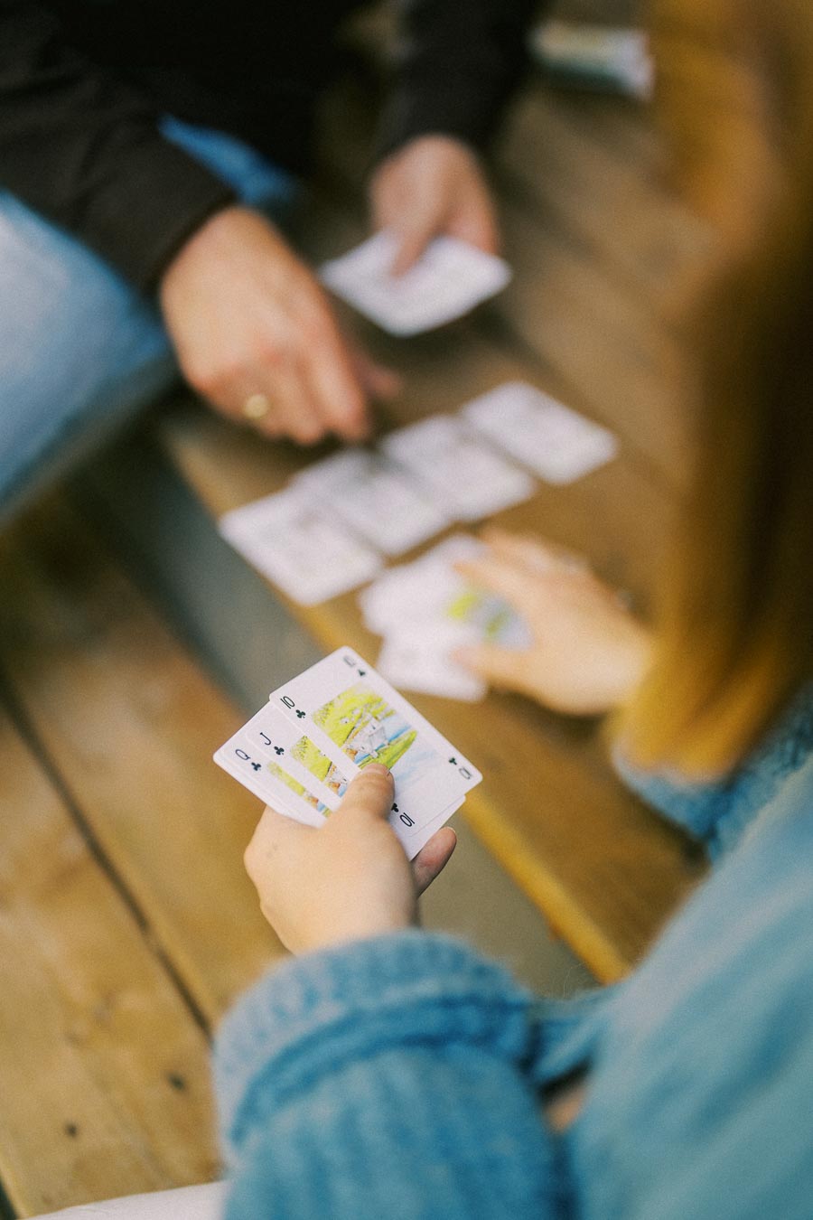 Two people playing a card game on wooden steps, focusing on one player's hand holding several playing cards close-up, emphasizing a social and leisurely activity.