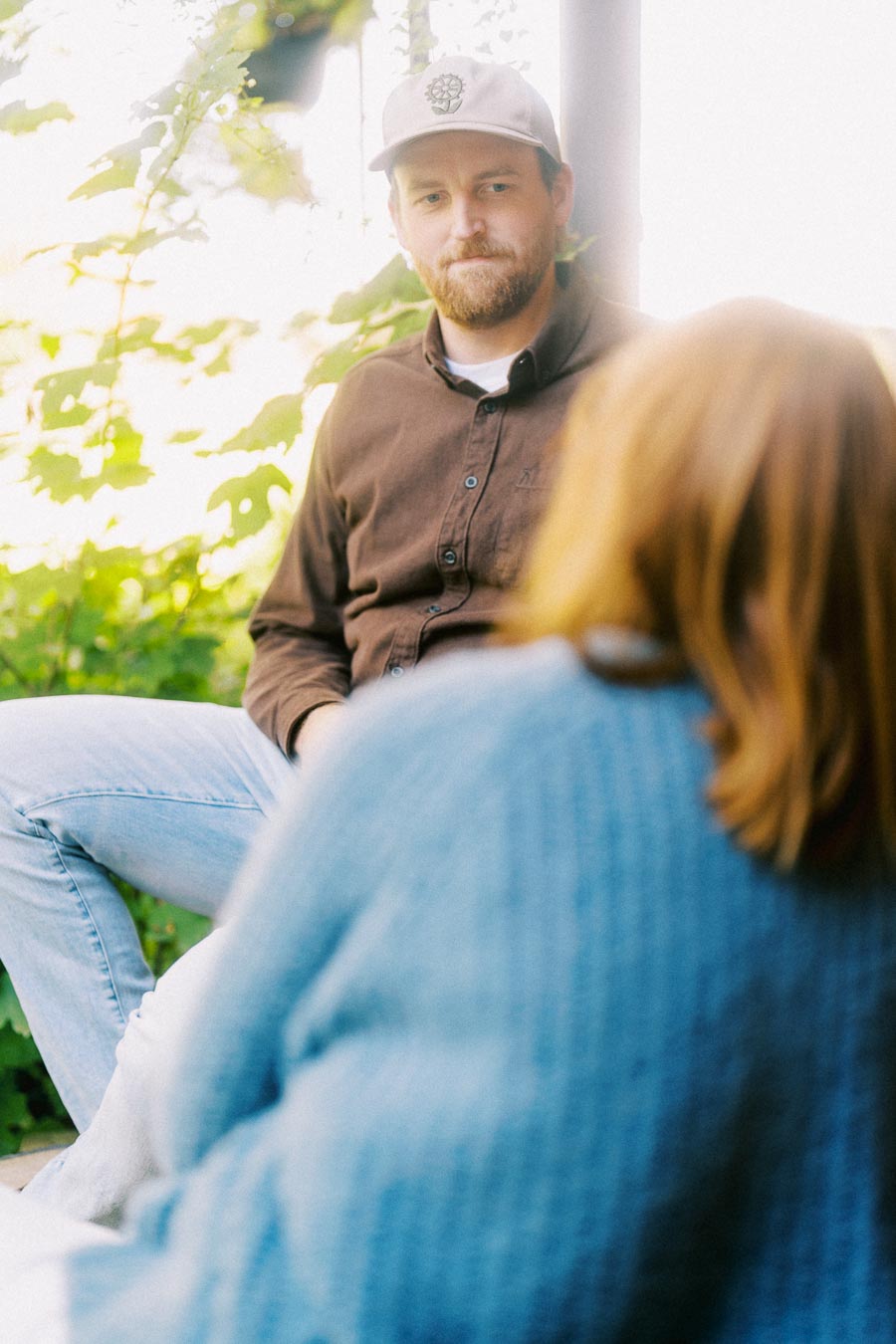 A man in a brown shirt and cap sitting outdoors, engaged in conversation, with blurred greenery in the background and a person in a blue sweater in the foreground.