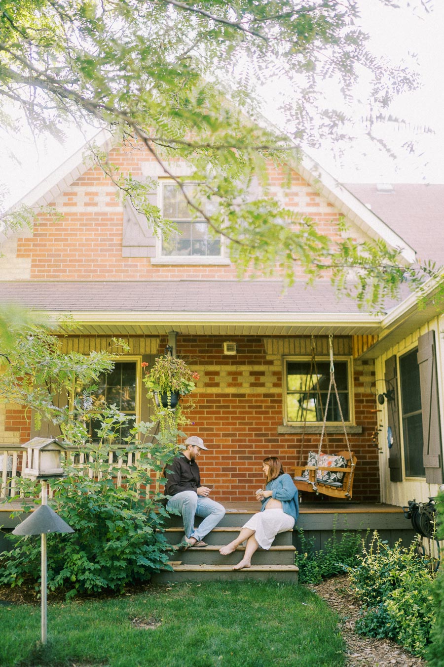 Cozy front porch of a red brick house featuring a couple sitting on steps, surrounded by greenery and a wooden swing chair, embodying a peaceful and inviting atmosphere.