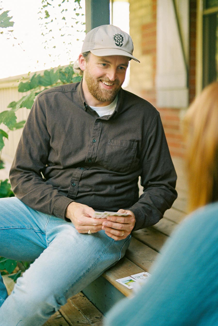 Man in a casual brown shirt and hat with a beard playing cards on a porch, smiling during a relaxed outdoor gathering.