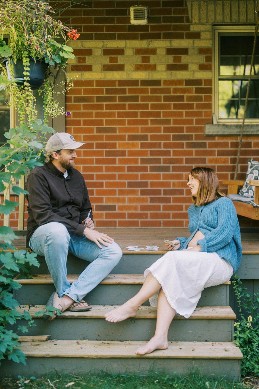 Couple enjoying outdoor conversation on porch steps, surrounded by lush greenery and a brick wall backdrop.