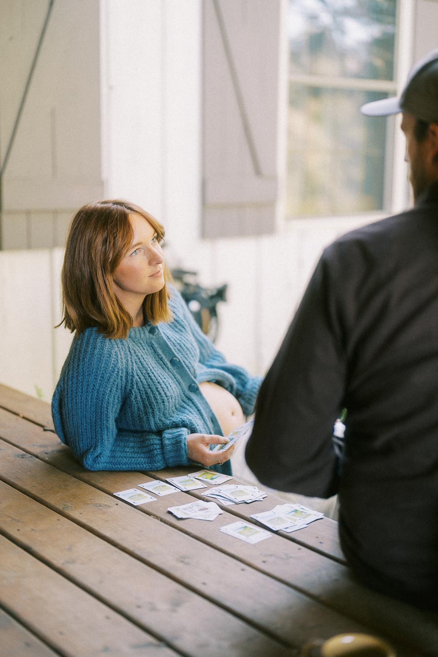 Pregnant woman in a blue sweater sitting at a wooden table, playing card game with another person.