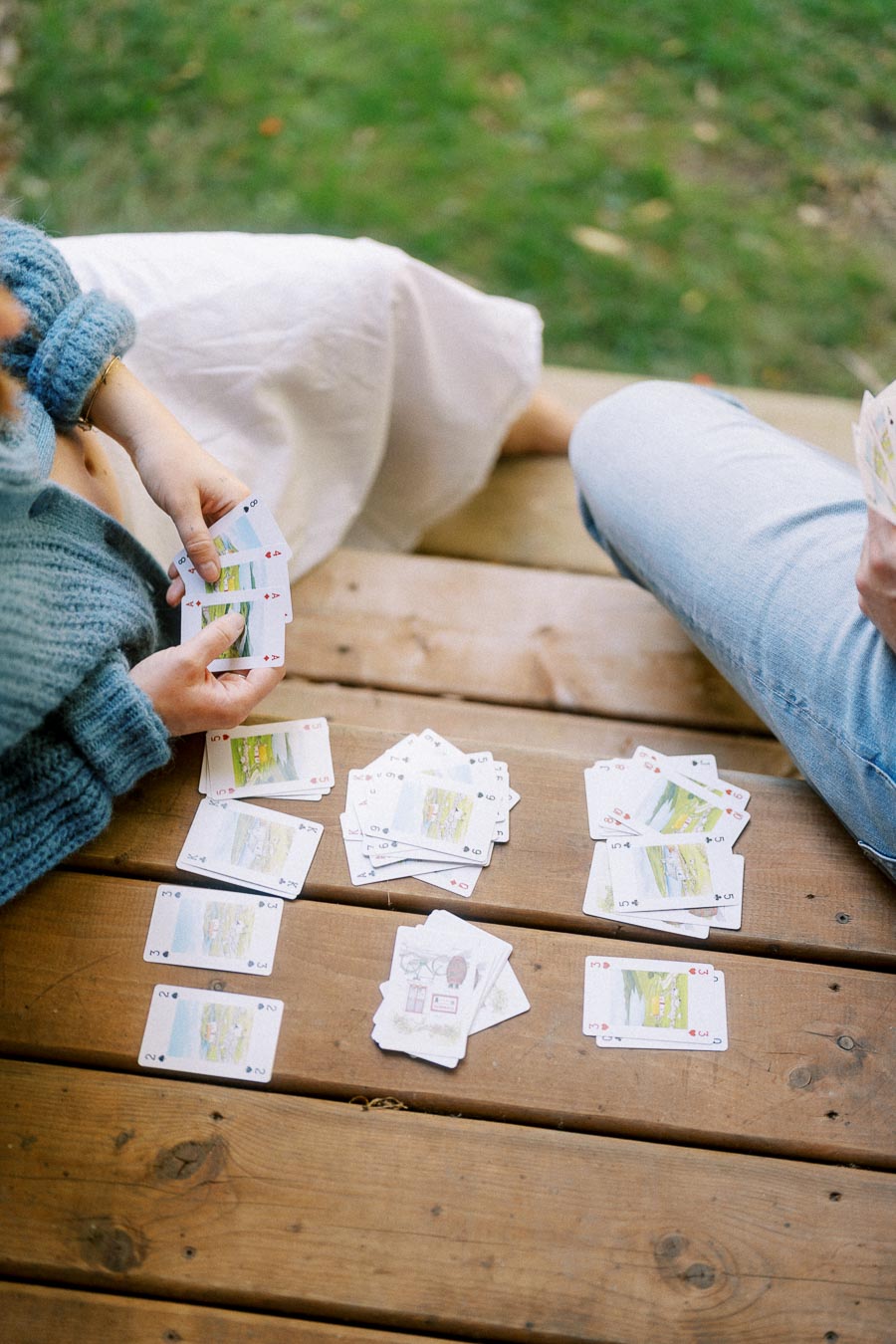 Two people playing cards on a wooden deck, surrounded by scattered playing cards.