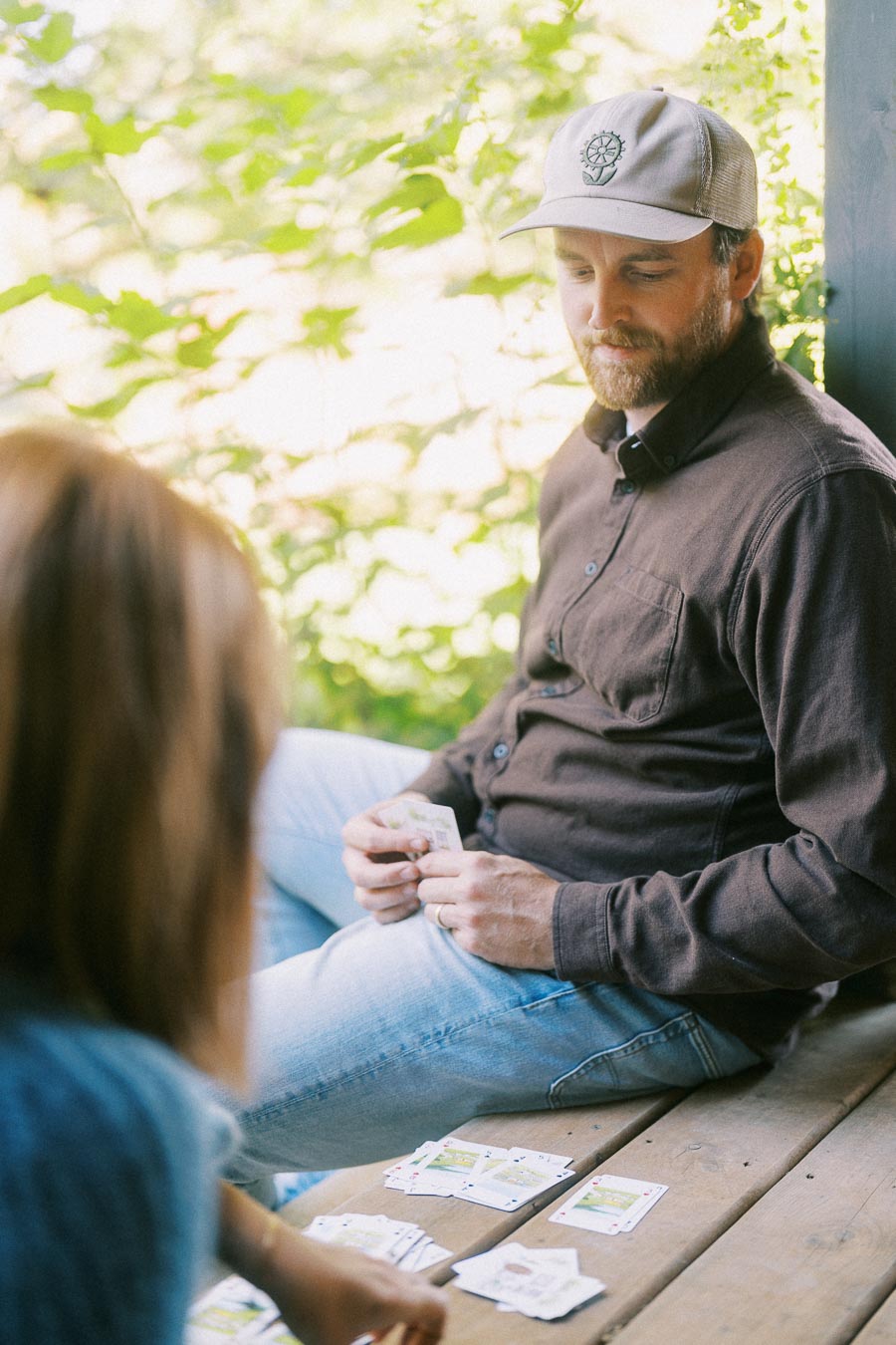 A man in a brown shirt and cap playing a card game with a woman on a wooden deck, surrounded by lush green foliage.