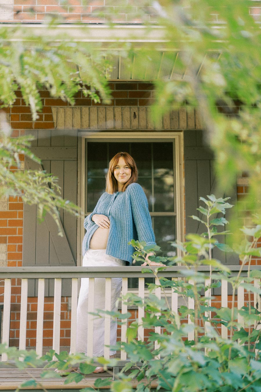 Pregnant woman in a blue sweater standing on a porch surrounded by lush greenery, smiling gently as she holds her belly, framed by a brick wall and wooden railings.