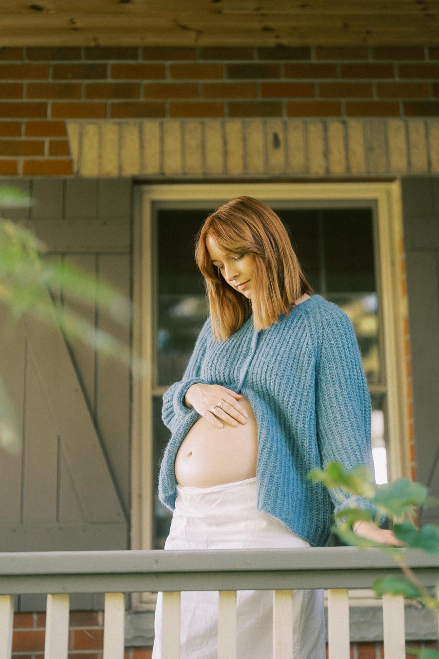 Pregnant woman standing on porch, wearing a cozy blue sweater and gently holding her belly.