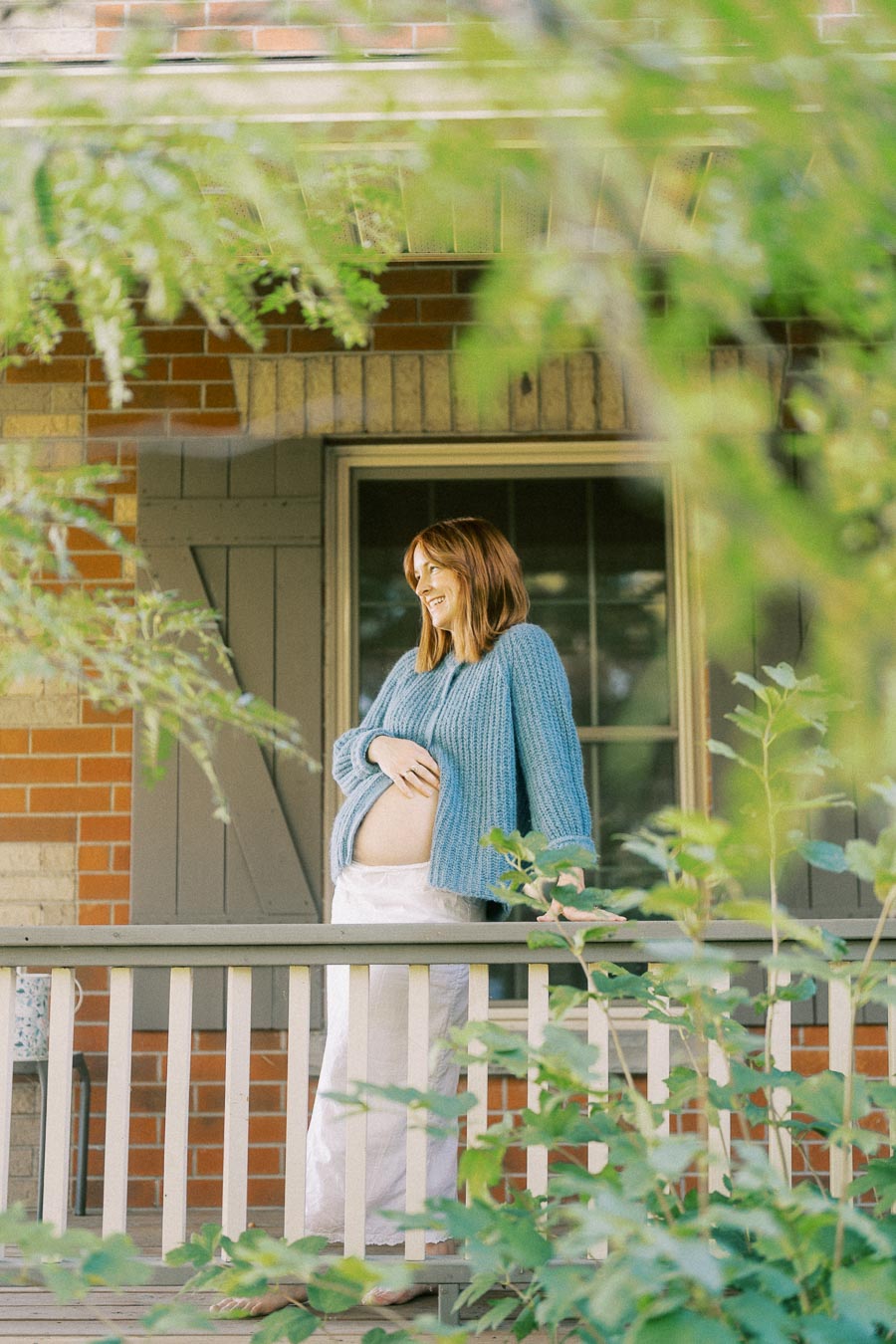 A pregnant woman wearing a blue sweater smiles while standing on a porch surrounded by greenery.