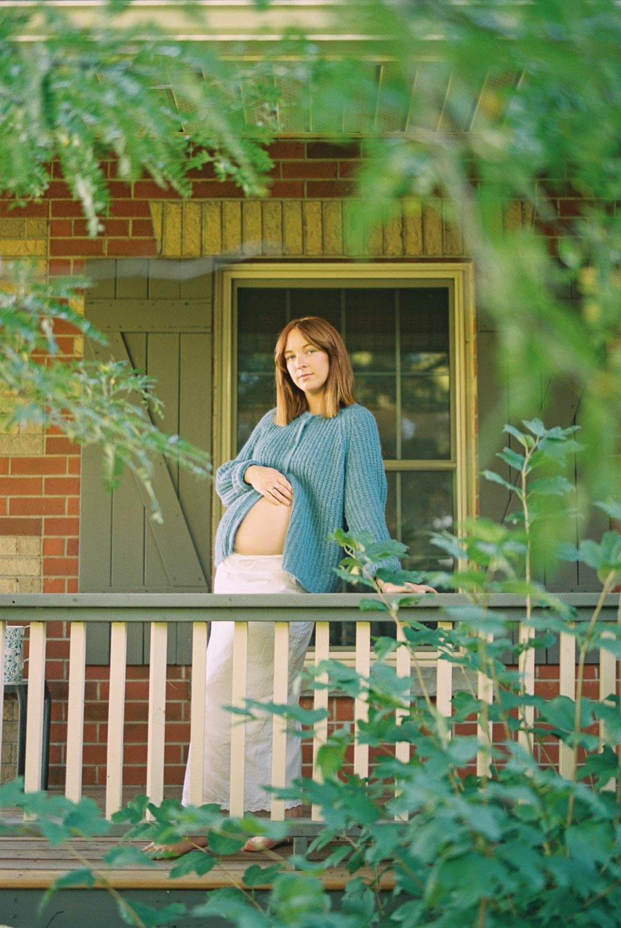 Pregnant woman in a blue sweater standing on a porch, surrounded by greenery, with a peaceful expression and hand gently touching her belly.