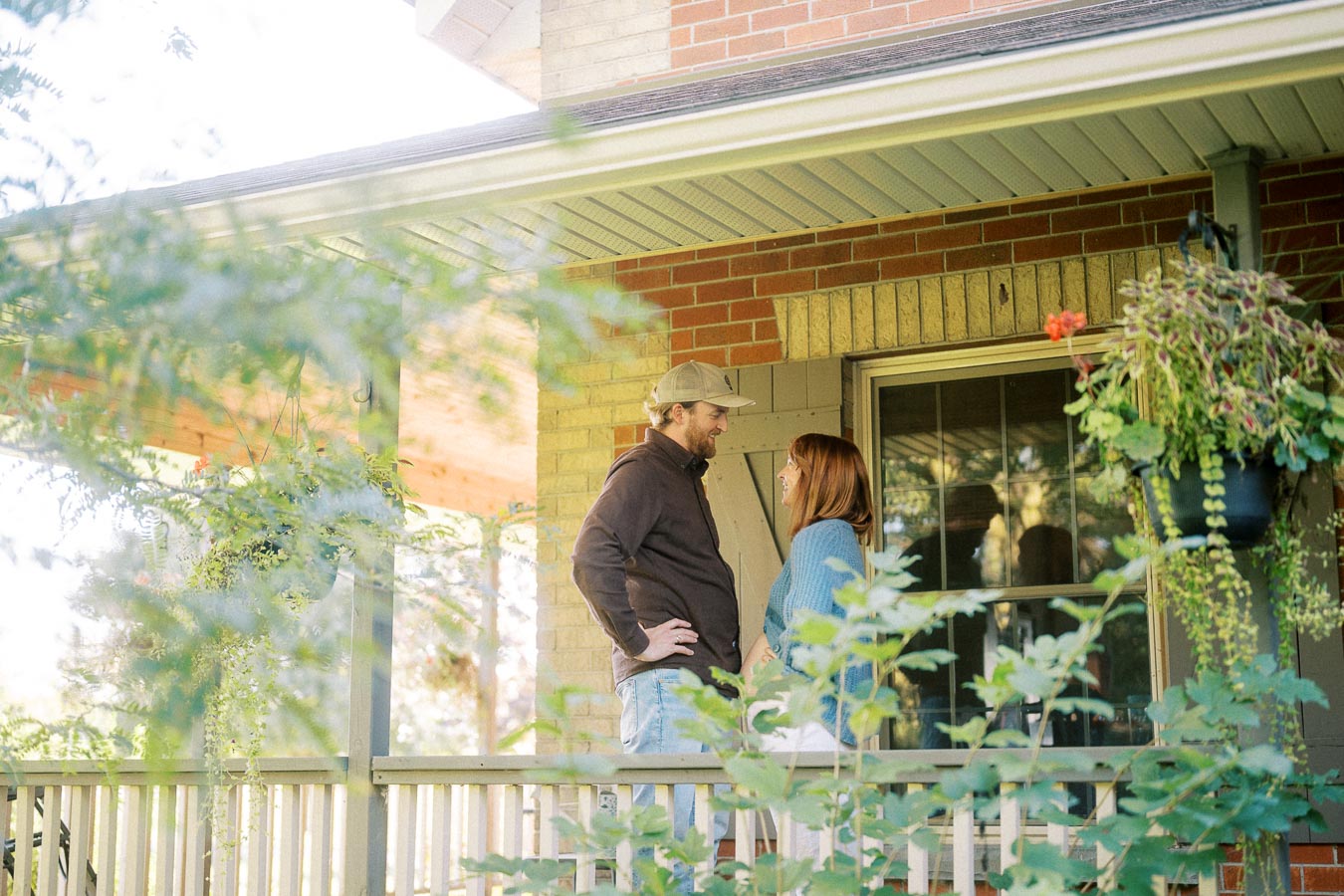 A man and woman smiling at each other on a porch surrounded by greenery, with a hanging flower pot nearby.