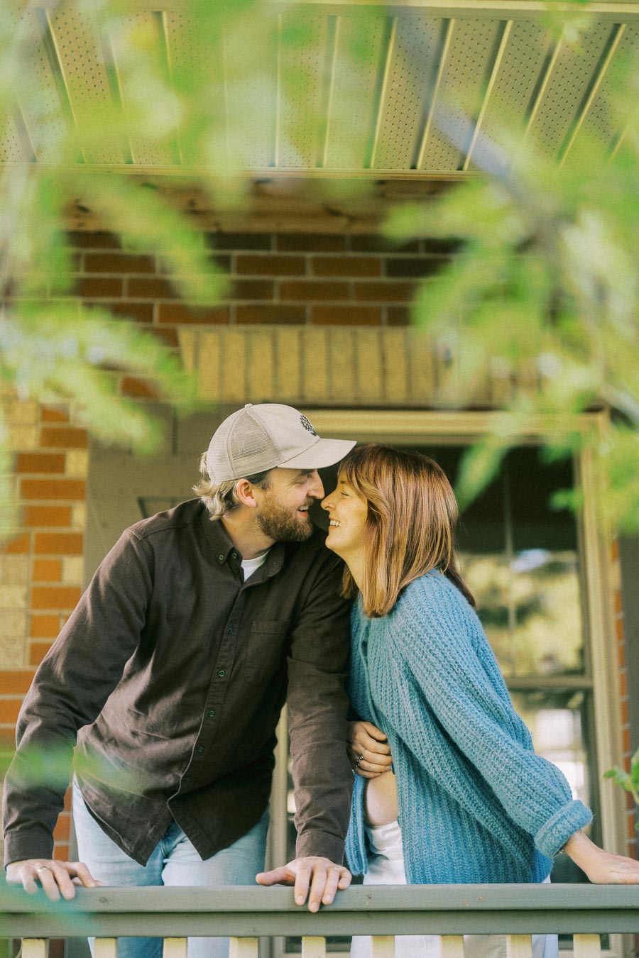 A couple sharing a loving moment on a porch, surrounded by greenery; the man wearing a cap and dark shirt, and the woman in a blue sweater, showing affection against a brick wall backdrop.