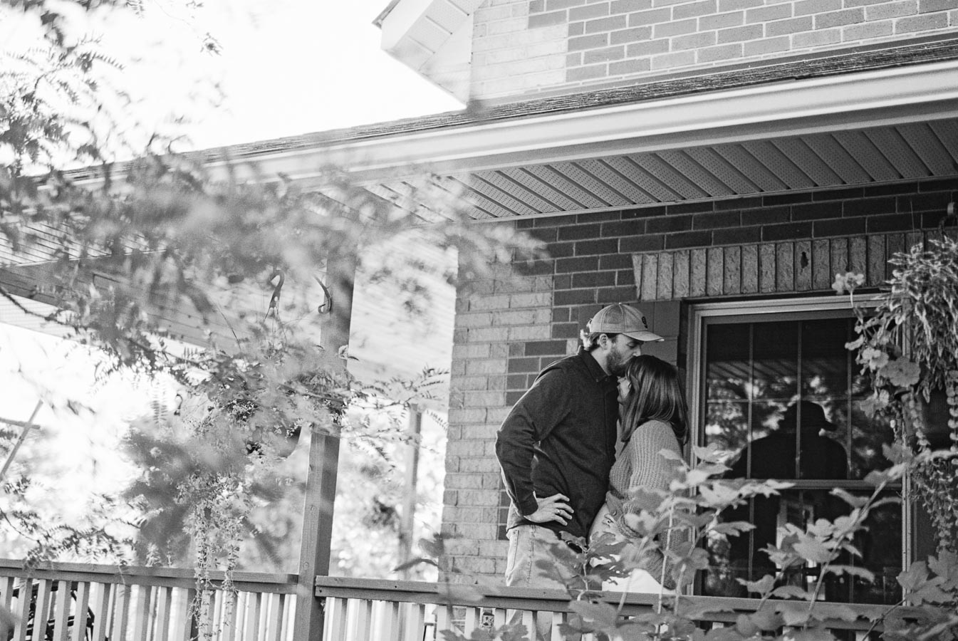 A couple embraces on a porch, surrounded by greenery and a brick house exterior.