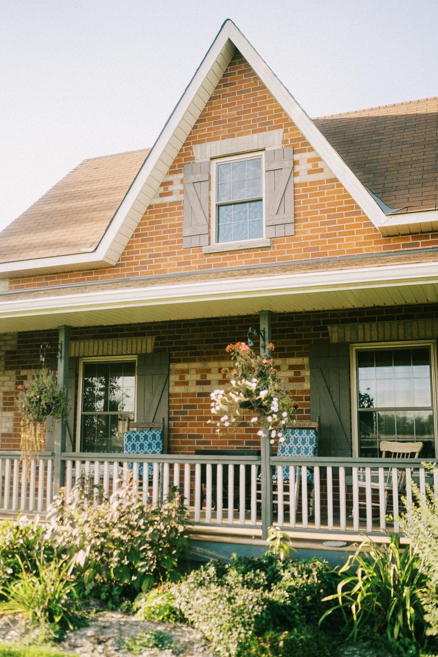 Charming brick house with a gabled roof and a cozy front porch surrounded by green landscaping, featuring hanging plants and outdoor seating.