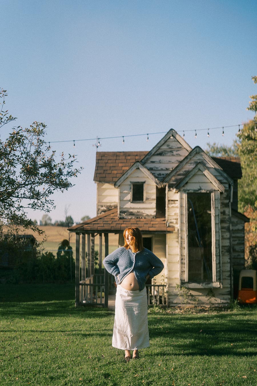 Pregnant woman in a rural setting, standing confidently in front of a rustic wooden house, wearing a blue sweater and white skirt, with string lights and clear blue sky in the background.